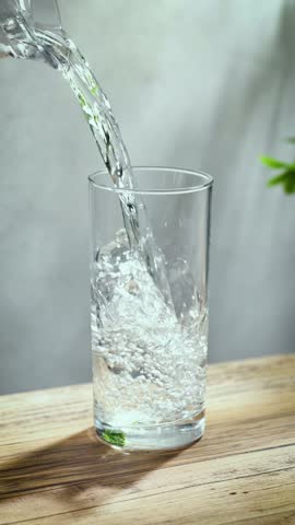 fresh clean water pouring into a glass on sunny wooden kitchen tabletop and gray wall background, slow motion
