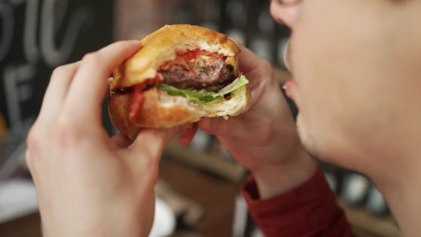 Young man wearing glasses savoring delicious burger at restaurant, enjoying every flavorful bite with evident pleasure