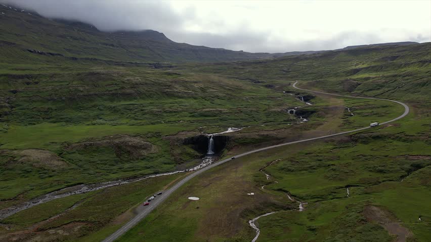 Stunning aerial footage of Seyðisfjörður Valley in Iceland. Featuring cascading waterfalls, a winding road, green landscapes, and mountain backdrops under a cloudy sky.