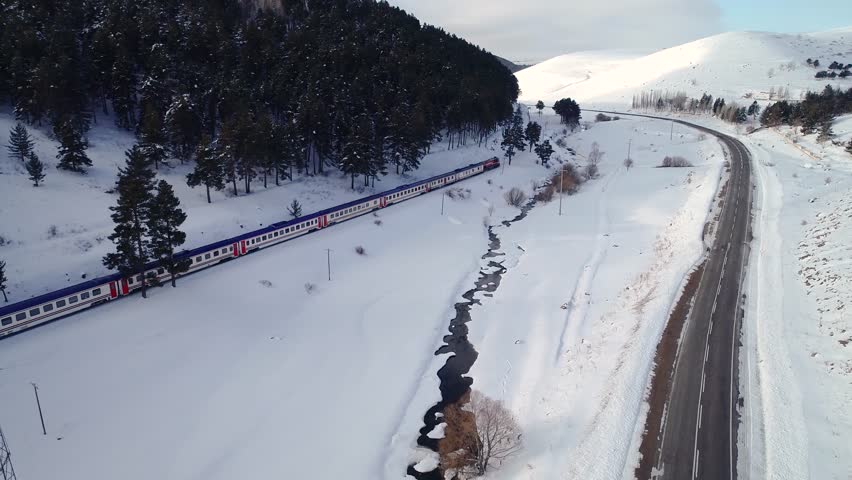 Aerial view of a train moving through a snow-covered forested valley. The winding tracks offer a peaceful journey through nature, blending transportation with the serene beauty of winter.