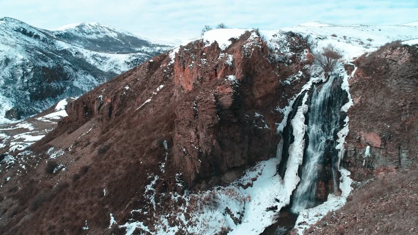A waterfall flows through snow-covered rocky mountains, creating a stunning winter landscape. The frozen rocks and cascading clear water capture the mesmerizing beauty of nature.