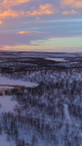 AERIAL: Flying above meandering river flowing through snowy winter wonderland in Lapland wilderness at dreamy sunset. Stream meandering through picturesque hilly countryside at golden sunrise, Finland