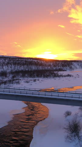 AERIAL: Flying above semi truck driving along scenic countryside highway at sunset. Transporter lorry crossing a bridge above frozen icy river in picturesque snowy landscape at gorgeous golden sunrise