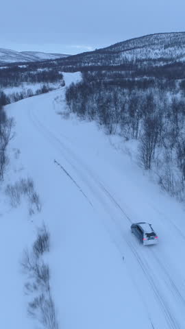 AERIAL: Flying above a car driving on scenic countryside highway at the bottom of forested snowy mountain. People on road trip across Lapland, Finland. Automobile traveling through wintry wilderness