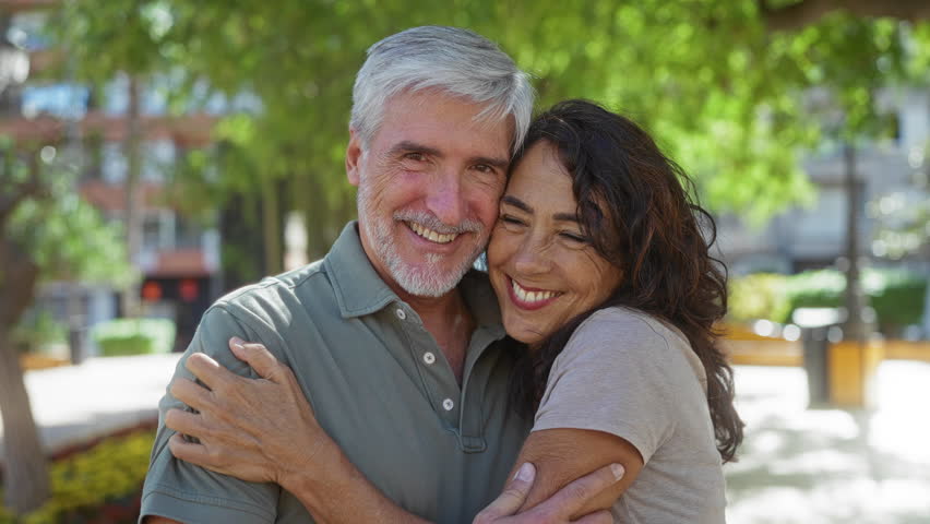 Middle-aged couple embraces outdoors in sunny park, showcasing love and happiness in an urban setting with smiling expressions.
