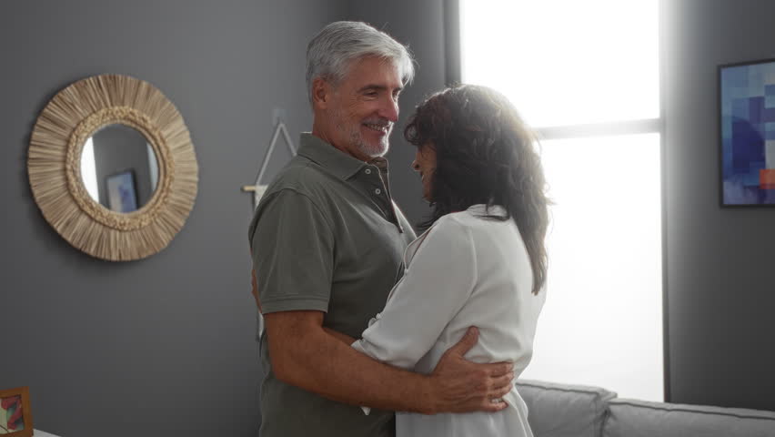 Middle-aged couple dancing happily in a cozy living room, the woman and man share a tender moment together in a warm home environment filled with love and intimacy.