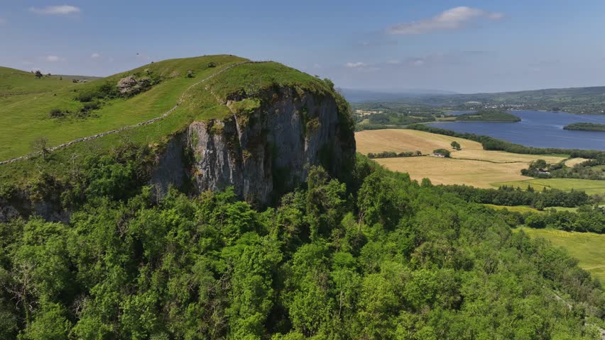 Hanging Rock, County Fermanagh, Northern Ireland. June 2023. Drone pushes forwards along the face of the cliffs above dense green woodland with Lough Macnean Lower in the background.