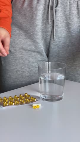 There are diverse vitamins on the table. A young woman is taking vitamins in turns and drinking them with a glass of water. Self-care, hydration, healthy lifestyle, and immune support are showcased.