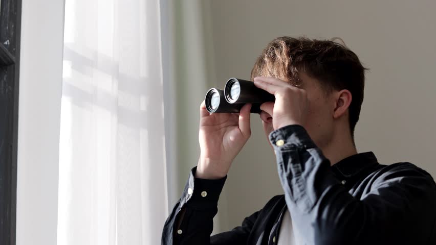 Young man stalker watching through binoculars in front of a window. Man spying or investigating.
