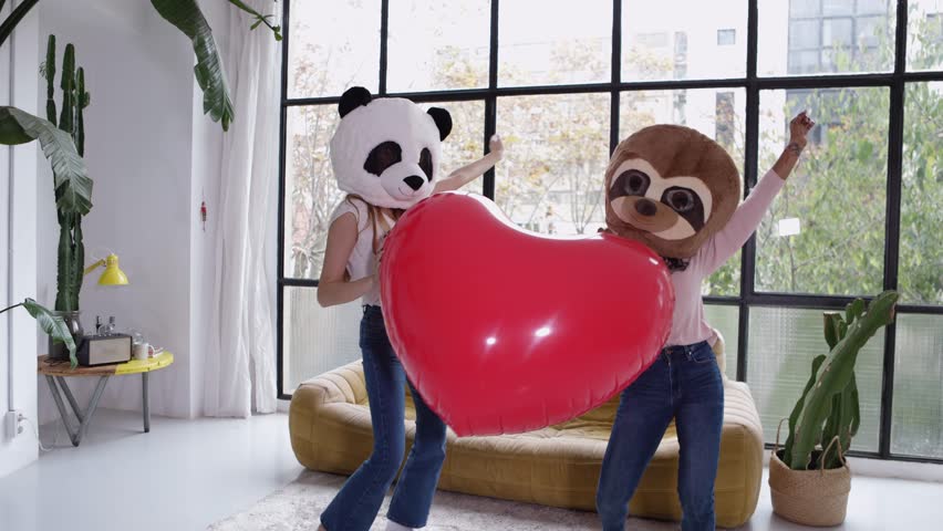 Two cheerful women wearing panda and sloth masks dancing while holding a big inflatable red heart and raising arms in a modern apartment