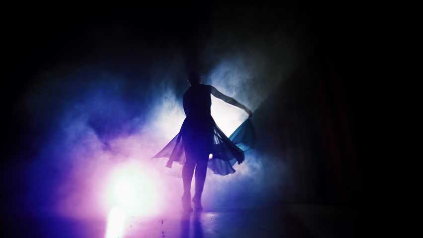 On a dark stage, a female dancer gracefully lifts her arms with a blue fabric. Stage lighting highlights her movement, while the curtain enhances the aesthetic and emotional depth of the performance.
