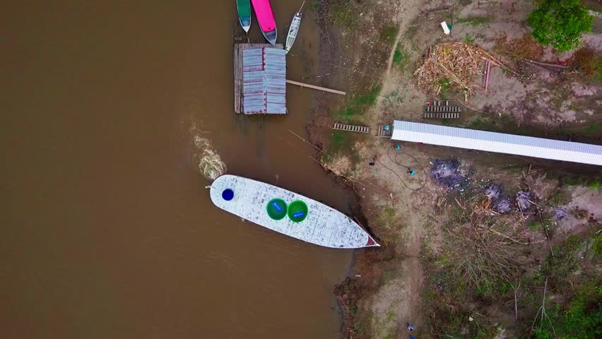 Rising top down aerial of boat by shore of brown Amazon River, Brazil