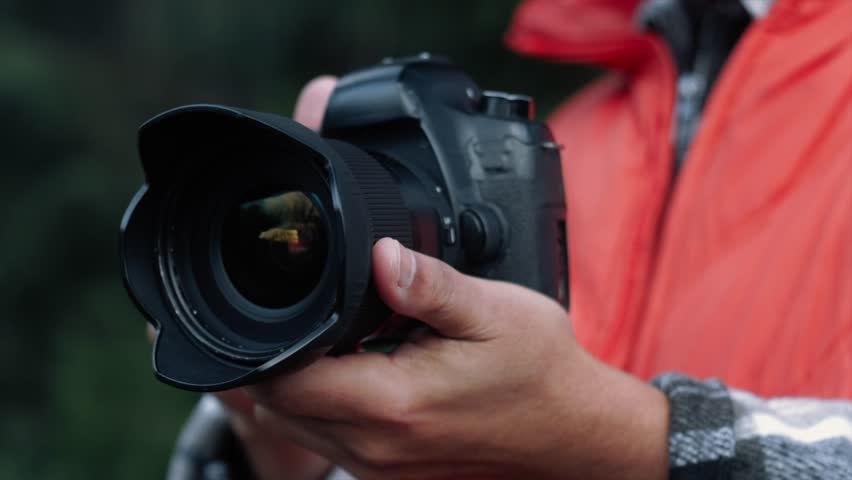 Professional photographer wearing bright orange jacket adjusting camera lens while standing outdoors, capturing detailed natural landscape with precision