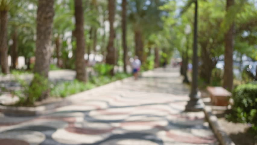 Blurred figures walking along a sunlit street in the palm tree-lined promenade of alicante, spain, featuring a colorful tiled walkway and lush greenery