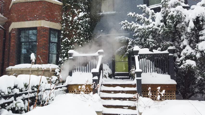 Snowy winter in Toronto, Ontario, Canada. View of a two-story brick house with a porch and patio covered in snow. Steam from the heating system rises from the basement of the house.
