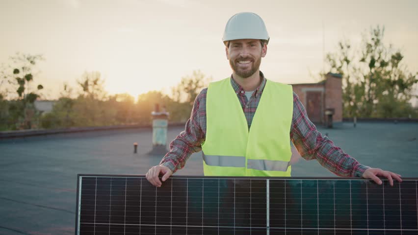 Engineer in reflective vest and helmet holding solar panel during sunset. Rooftop solar installation showcases dedication to renewable energy. Portrait of happy man with sustainable progress concept.