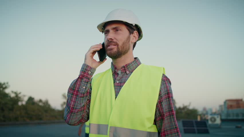 Project manager on call discusses solar panel installation updates, sustainable energy goals. Caucasian young man in helmet talking on mobile phone. Male worker speaking on telephone outdoor.
