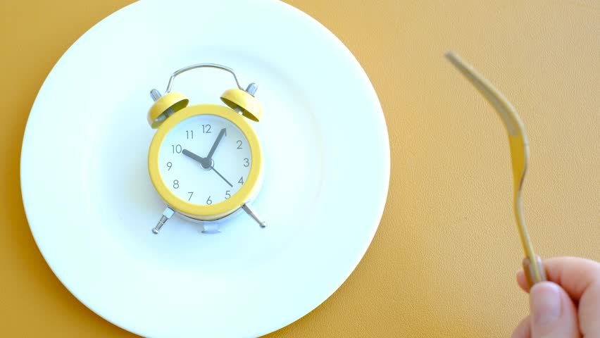 Empty plate with alarm clock and fork on brown background, intermittent fasting concept.