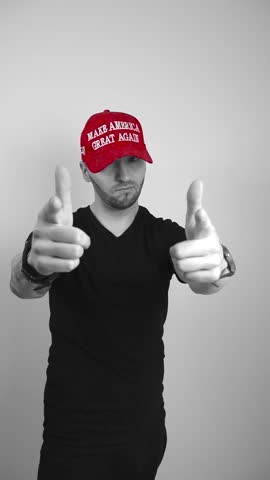 Grayscale footage of a man with tattoos who is wearing a red colored MAGA Make America Great Again Trump presidential hat lifting up his arms and points at the camera while nodding with happy approval