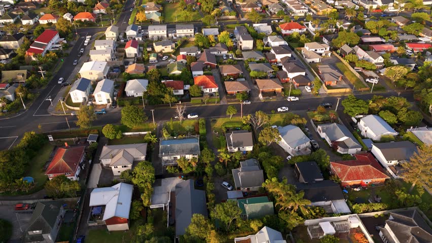 Suburb neighborhood with single Family house and parking cars on street during golden sunset. Waterview, District in Auckland, New Zealand. Downtown skyline in distance Aerial tilt up wide shot.