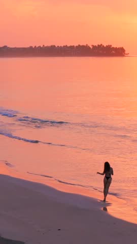 Vertical Pretty beautiful slender model woman in swimsuit running by the edge of water on the wild tropical sandy beach at amazing pink sunset. Romantic woman enjoying life and having fun 