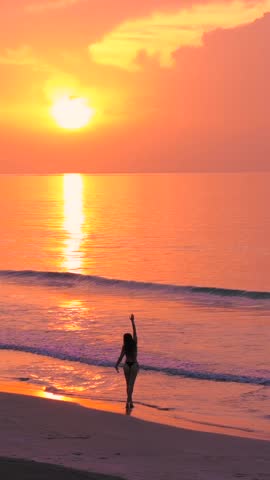 Vertical silhouette of a woman in a swimsuit, jogging along the beachfront of a remote tropical island at a majestic pink sunset.
