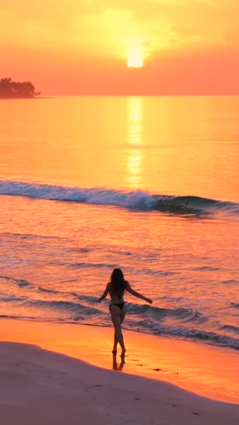 Vertical silhouette of a pretty beautiful slender model girl in swimsuit running by the edge of water on the beach at amazing pink sunset.