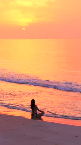 The silhouette of a young slender woman in yoga meditation pose sitting on a wild tropical beach with no people at beautiful sunset. Relaxation and calm to relieve stress and anxiety