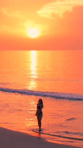 Vertical silhouette of girl in swimsuit walking, spinning and dancing on a wild tropical sandy beach at amazing pink sunset. Romantic woman enjoying life and having fun 