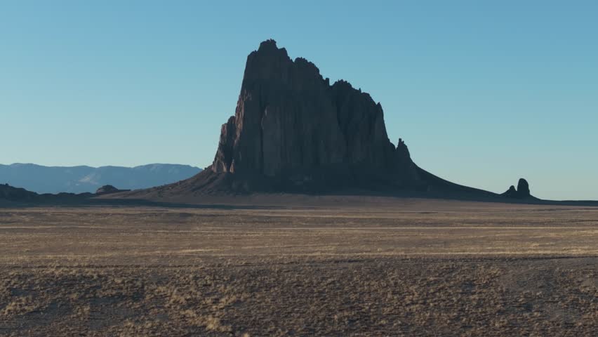A low to the ground, slow-rising 70mm drone shot of the sacred “Ship Rock”, a massive, monolithic monadnock towering above the high-desert plain of the Navajo Nation, in New Mexico, United States.