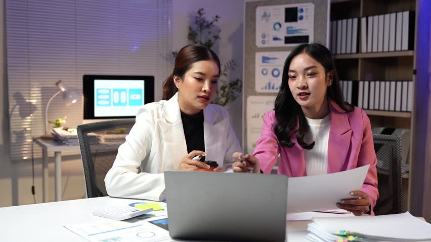 Two young Asian businesswomen are working late in office, analyzing financial data on laptop and documents, discussing and collaborating on project
