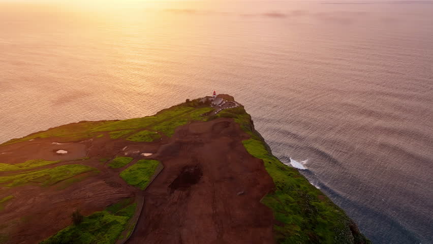 Stunning aerial view of a coastal lighthouse at sunset overlooking the ocean waves and green hills