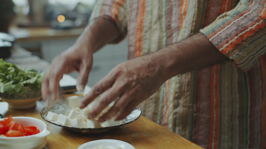Hands of unrecognizable man coating cubes of fresh paneer cheese with flour in plate at wooden kitchen countertop with chopped tomatoes, onions and fresh herbs on it. Close-up view