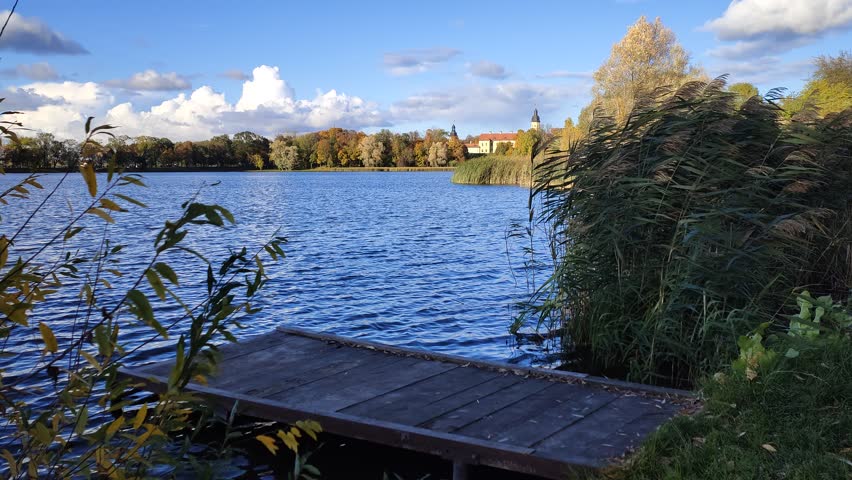 In the evening, the autumn sun sinks to the horizon and illuminates the medieval castle and the trees with yellow leaves. A wooden pier and reeds stand near the grassy bank. Blue sky with clouds