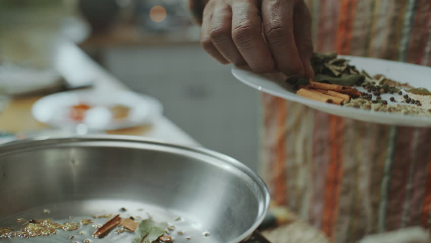 Hands of male chef adding whole spices like bay leaves, cinnamon sticks and peppercorns to heated pan for cooking Indian food in kitchen. Close-up shot