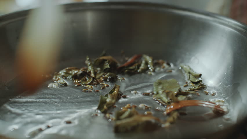 Close-up view of unrecognizable chef using spatula to stir whole Indian spices such as bay leaves and cumin seeds in hot oil inside frying pan, releasing aroma and flavor for cooking