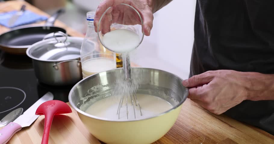 Man adds sugar to liquid crepe dough in metal bowl on table. Male cooks homemade pancakes of natural ingredients in light home kitchen slow motion