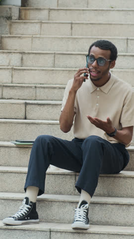 Vertical shot of cheerful African American male entrepreneur talking on smartphone sitting on urban stairs