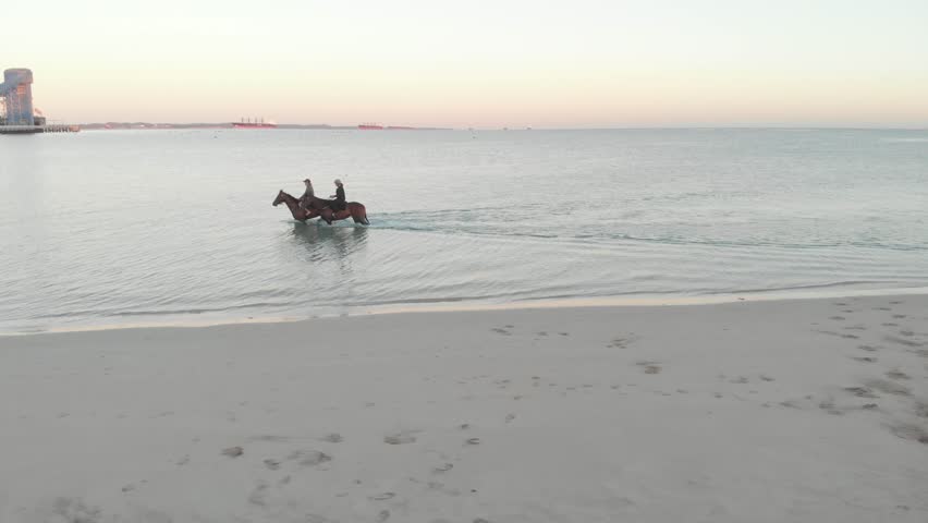 A drone scenic view of two persons riding horses in the sea at sunset in Rockingham, Australia