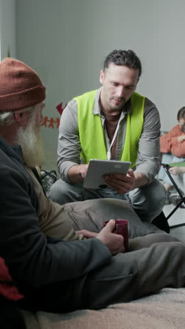 Full vertical shot of attentive Caucasian male emergency responder in yellow vest sitting next to senior male refugee with beard, asking questions, filling checklist on digital tablet