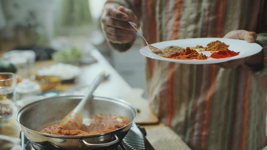 Hand of chef sprinkling aromatic spices from plate into simmering tomato sauce in pan on stove, cooking traditional Indian dish in kitchen. Close-up shot