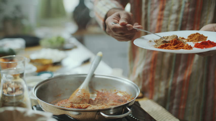 Hand of chef sprinkling aromatic spices from plate into simmering tomato sauce in pan on stove, cooking traditional Indian dish in kitchen. Close-up shot