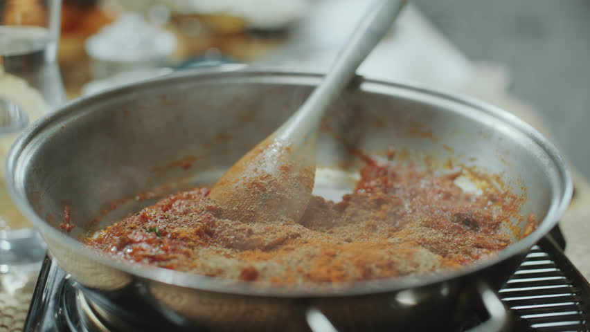 Hand of man using spoon to add aromatic Indian spices to steaming tomato sauce in pan, cooking traditional dish in kitchen. Close-up shot