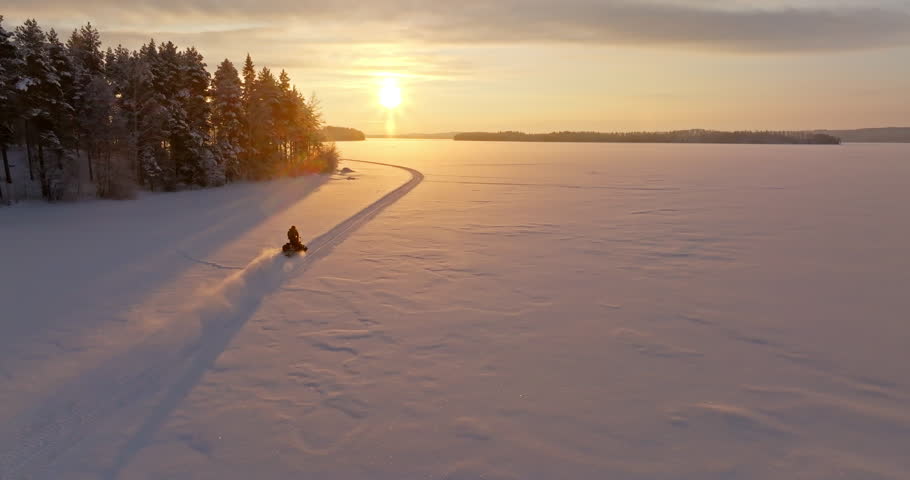 Drone shot following a snowmobile driving on ice, toward the sunset of Lapland