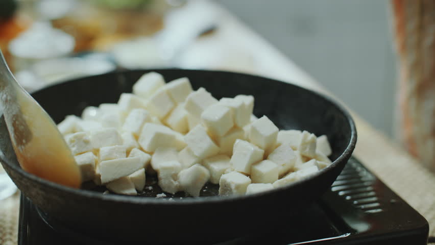 Hand of chef stirring cubes of fresh paneer in hot frying pan with spatula, cooking traditional Indian food in kitchen. Close-up view