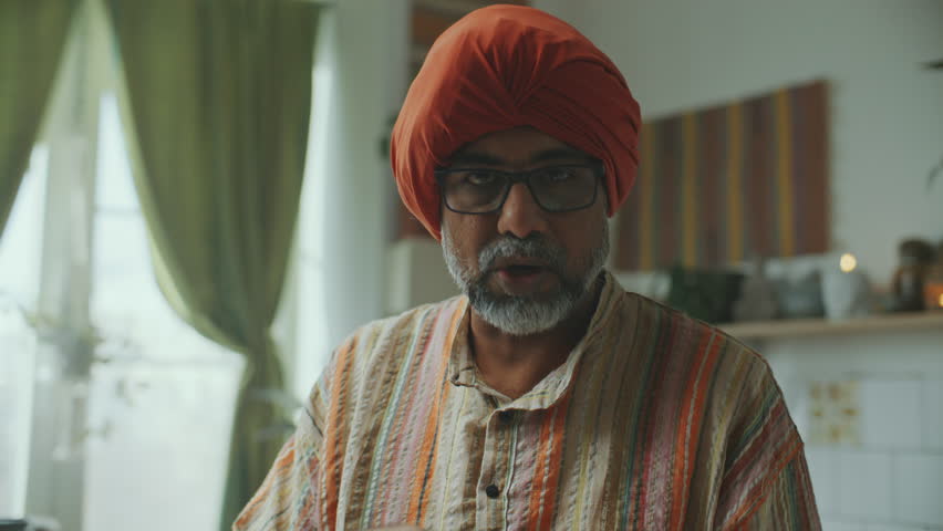 Indian food blogger in turban talking to camera, then adding cubes of paneer to rich spiced tomato sauce in pan on stove, stirring ingredients together as preparing traditional dish in kitchen