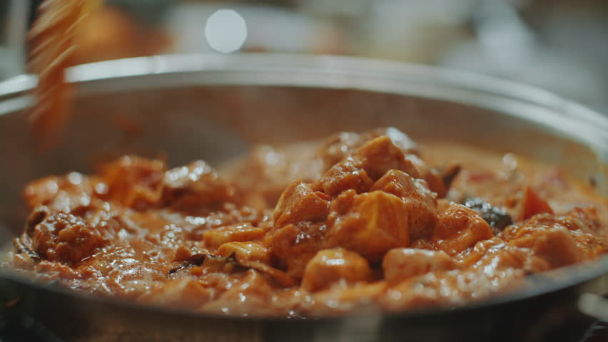 Extreme close-up shot of hand stirring creamy paneer butter masala in pan, mixing ingredients during cooking process of flavorful Indian dish