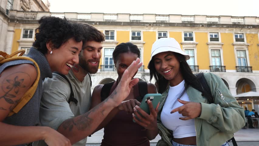 Diverse group of young travelers using smartphone for navigation, sharing joyful moments while exploring historic comercio square in lisbon, portugal during sunny summer day