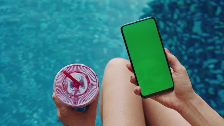 Woman relaxes holding smartphone with green screen and refreshing drink by pool closeup. Lady enjoys summer vacation at luxury resort. Mobile devise with chromakey screen