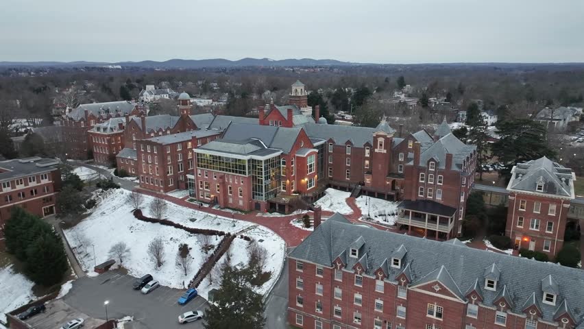 Aerial approaching shot of historic private Randolph College School in Lynchburg, Virginia,USA. Snowy winter day in America. Leafless forest trees in distance.